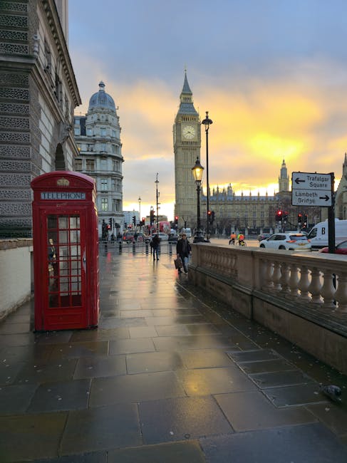 A wide sidewalk along a street in Lambeth, London, during sunset with cobblestone paving, a traditional red telephone box on the left, and historic buildings with ornate stonework. Street lamps are lit, and pedestrians walk along the promenade. In the background, Big Ben and the Houses of Parliament stand prominently under a partly cloudy sky with warm orange and blue hues. The scene captures an iconic view of central London, relevant to local urban environments, though there are no surfaces or cleaning elements visible related to residential or commercial cleaning processes.