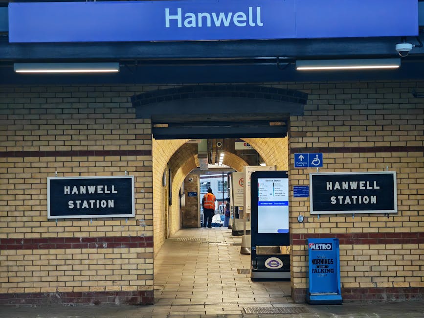 The entrance to Hanwell Station features a brick archway with two black and white signboards labeled 'Hanwell Station' on either side. Above, a blue sign displays the station name 'Hanwell' in white letters. The passageway beyond the arch reveals a tiled walkway with a person in an orange safety vest walking inside. On the right side, there is a digital ticket machine and a blue Metro timetable display, while a wheelchair accessible sign is mounted on the wall. Bright fluorescent lighting illuminates the open corridor, and the overall scene appears clean and well-maintained, emphasizing the station's orderly environment.