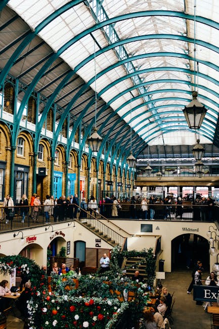 Interior view of Brixton Market showcasing a high, arched glass ceiling supported by teal-colored metal framework, allowing natural light to illuminate the space. The market floor features a mixture of brick and tiled surfaces, with vendors' stalls and seating areas visible on multiple levels. Customers and visitors are seen walking and sitting, with some engaged in conversations or browsing. Potted plants and flower arrangements add color and vibrancy to the bustling environment. The overall scene depicts a clean and well-maintained space, emphasizing hygiene and order consistent with professional commercial cleaning standards. Lambeth Carpet Cleaning offers surface cleaning and sanitisation services suitable for busy public venues like Brixton Market, ensuring high standards of cleanliness and hygiene.