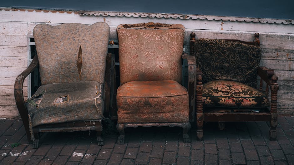An outdoor scene featuring three antique upholstered armchairs arranged in a row against a weathered wooden wall. The left chair has a torn, faded fabric with exposed cushioning and frayed edges, indicating significant wear. The middle chair displays a similar age-related fabric deterioration with a slightly faded floral pattern and visible stains. The right chair has a dark, patterned upholstery with a more intact appearance but shows signs of age and dust accumulation. All three chairs are positioned on a brick-paved surface, with natural light illuminating the scene, highlighting their textures and worn condition. Lambeth Carpet Cleaning emphasizes thorough surface and upholstery cleaning, even for aged furniture, as part of their domestic cleaning services.