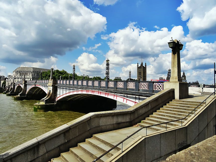 Photograph of the Westminster Bridge crossing the River Thames in London, with ornate lamp posts along the bridge railing and stone stairs leading down to the riverbank in the foreground. The scene is illuminated by natural daylight, highlighting the bridge's white and pink detailing, the calm water beneath, and the historic Gothic architecture of the Palace of Westminster in the background, topped with a partly cloudy sky. This image captures an iconic urban landscape, emphasizing outdoor surface surfaces, materials like stone and metal, and the cleanliness and structural details of the bridge and surroundings. Lambeth Carpet Cleaning as a professional service promotes maintaining hygienic and well-maintained environments, akin to the importance of keeping such public surfaces clean and presentable.