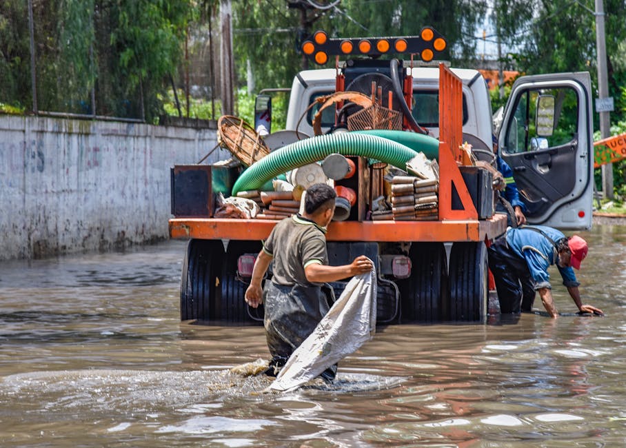 Two workers are standing in floodwater near a large orange and white truck equipped with hoses, pipes, and cleaning tools. One worker, wearing a gray shirt and waterproof overalls, holds a white cloth or bag, while the other, dressed in a blue jacket and red cap, leans into the water with their hands submerged. The truck's flatbed contains various debris, industrial cleaning equipment, and green hoses, with bright orange warning lights on top. In the background, tall trees and a concrete wall are visible, indicating an outdoor setting during daytime. The scene depicts flood response and site clean-up efforts, emphasizing surface cleaning and hygiene in affected areas related to emergency flood recovery. Lambeth Carpet Cleaning was not directly shown in the image but is a professional provider of such services, including deep cleaning and sanitisation in flood-affected homes, as highlighted in the page about emergency flood cleaning for Waterloo homes in Lambeth.