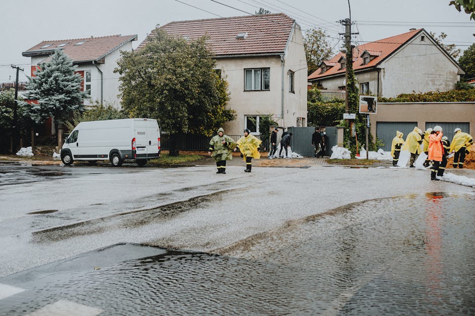 A residential street in Waterloo with wet asphalt reflecting the overcast sky, where a team of emergency flood cleaning workers in yellow and orange raincoats are managing a flood response near two white houses with tiled roofs. The workers are seen walking and standing on the damp roadside, with some near a fenced property and others by a pile of white sandbags along the sidewalk. A white commercial van is parked on the side of the street. The surroundings include trees and utility poles, with evidence of recent heavy rain and flooding, emphasizing the importance of surface cleaning and sanitisation services provided by Lambeth Carpet Cleaning for flood-affected homes.