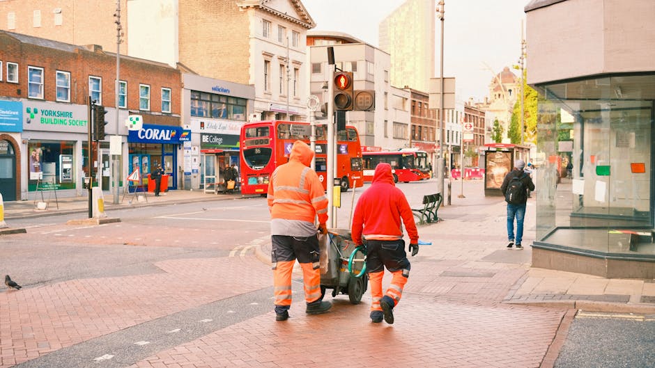 Two professional cleaning workers dressed in high-visibility orange and red uniforms are walking on a city street in Lambeth, London, pushing a cleaning trolley. They are surrounded by urban infrastructure, including shops such as Yorkshire Building Society and Coral, as well as buses and pedestrians. The scene is illuminated by daylight, and the street surface appears clean and well-maintained, reflecting a focus on surface cleaning and hygiene in a busy commercial area. Lambeth Carpet Cleaning emphasizes thorough professional cleaning in such environments, ensuring high standards of sanitation and surface upkeep.