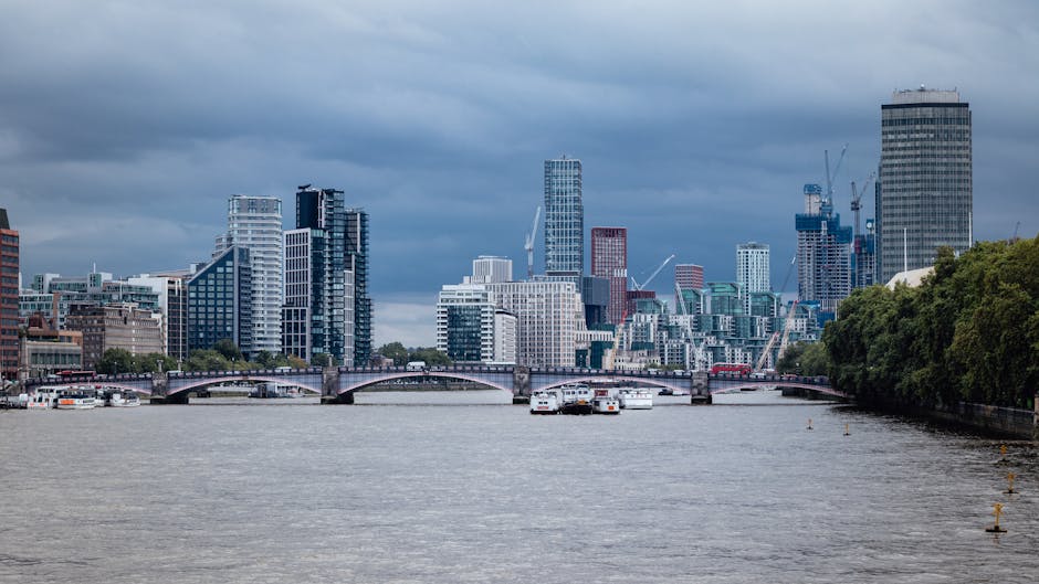 A panoramic view of the London city skyline over the River Thames, showcasing modern high-rise buildings with glass facades, some under construction with cranes, set against a cloudy sky. In the foreground, the river's surface appears calm with a few boats and vessels docked along the banks. To the right, lush green trees line the riverbank, adding natural contrast to the urban scene. The area is illuminated by natural daylight, highlighting the architectural details and reflecting off the water.