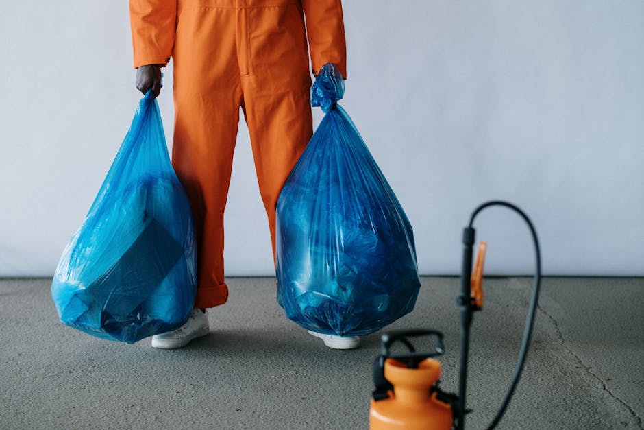 A person dressed in orange work overalls and grey gloves is standing on a smooth grey carpeted floor, holding two large blue plastic trash bags filled with waste. The setting appears to be an indoor space with a plain white wall in the background. Visible in the foreground is an orange cleaning spray bottle connected to a black spray nozzle with a hose, suggesting cleaning or waste disposal activities. The lighting is even and neutral, emphasizing the cleanliness and organization typical of professional surface cleaning and waste management. The image reflects a professional cleaning service environment, in which waste collection and disposal are part of routine hygiene and maintenance practices, consistent with lambethcarpetcleaning.com's focus on cleaning and sanitation.
