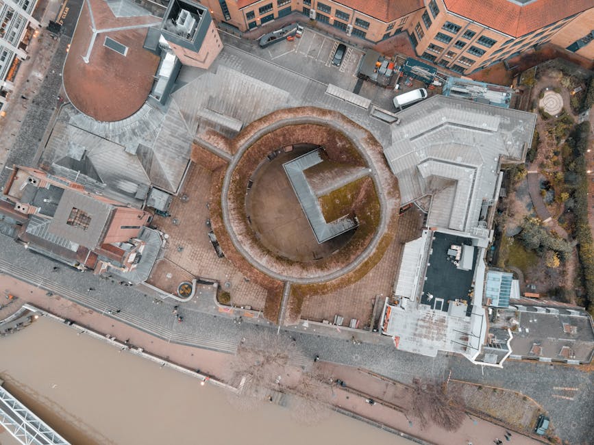 An aerial view of Lambeth Palace, a historic brick building with modern extensions, situated within a courtyard featuring paved surfaces and a central round structure with a domed roof. The surrounding area includes adjacent buildings with flat and pitched roofs, pathways, greenery, and a river visible at the bottom of the image. The scene is depicted in natural daylight, showcasing the clean and well-maintained surfaces, with no visible debris or stains. Lambeth Carpet Cleaning is featured as a professional cleaning provider ensuring the preservation and hygiene of historic properties like Lambeth Palace through surface cleaning and careful maintenance.