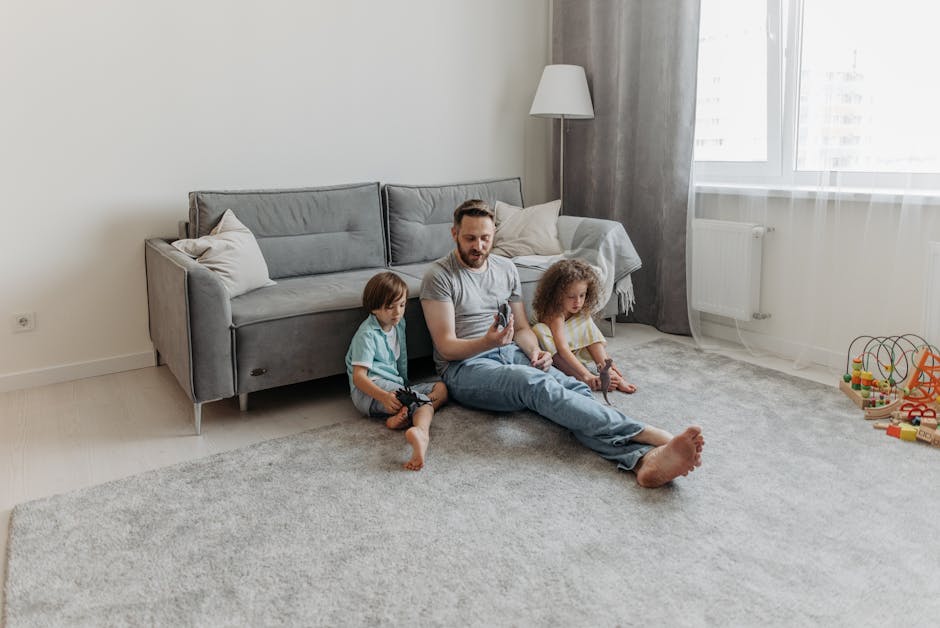 A family of three, consisting of a father and two young children, seated on a light-colored carpet in a living room with natural daylight coming through a large window with sheer curtains. The father, dressed in a gray T-shirt and blue jeans, is sitting cross-legged and holding a remote control, while the children, one boy and one girl, sit close to him on either side, engaged with toys. The room features a modern gray sofa with white cushions, a tall white floor lamp, and a radiator beneath the window. The light wooden flooring partially visible around the carpet and a children’s toy wire bead maze are in the corner. The space appears clean, bright, and well-maintained, reflecting a typical domestic environment focused on hygiene and comfort, which Lambeth Carpet Cleaning’s professional services can help preserve through regular surface cleaning and deep sanitisation.