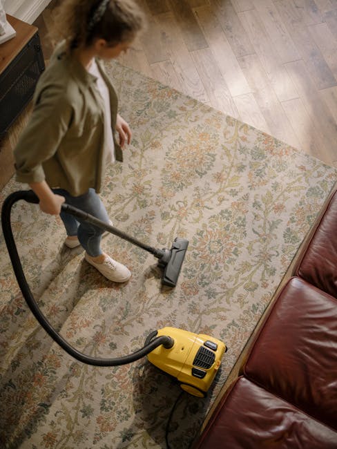 A woman using a yellow vacuum cleaner with a long hose on a large, patterned area rug in a living room. The rug features intricate floral designs in muted tones and covers a wooden floor. She is dressed casually in a beige jacket, jeans, and white sneakers. The room is well-lit, and part of a red leather sofa is visible on the right side. Lambeth Carpet Cleaning offers professional surface cleaning and deep cleaning services, maintaining hygiene and cleanliness in residential spaces, as seen in this domestic cleaning scene.