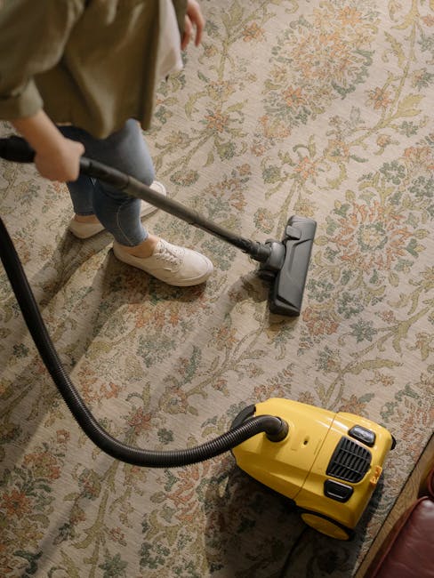 A person kneeling on a patterned, woven carpet in a room with natural lighting, using a yellow and black vacuum cleaner, possibly a wet-dry model, to perform surface cleaning. The individual is holding a white cloth or filter and placing it into the open top compartment of the vacuum. The carpet appears clean and well-maintained, with intricate floral and geometric designs, and the surrounding area is free of dust or debris. This scene demonstrates routine domestic cleaning and maintenance, reflecting professional standards provided by Lambeth Carpet Cleaning for residential or commercial spaces, in line with the focus of the page about same-day carpet cleaning in Lambeth for optimal hygiene and surface sanitisation.