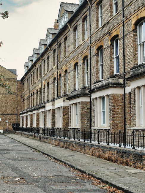 Photograph of a multi-story Victorian-style residential building with brick facade and white window frames, situated along a paved street in Lambeth. The building features several evenly spaced windows with small white sills, and a wrought iron fence runs parallel to the sidewalk at the base. The pavement appears clean, with some fallen leaves scattered, and the overall scene is illuminated by natural daylight. The image is relevant to interior deep cleaning services provided by Lambeth Carpet Cleaning for flats in the SE11 area, focusing on surface cleaning, hygiene, and maintenance of residential properties.